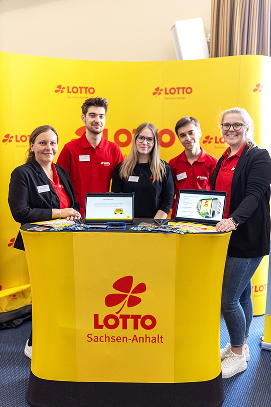 Das Messe-Team von LOTTO Sachsen-Anhalt auf der Berufsfindungsmesse der IHK in Magdeburg- Sie stehem am gelben Messestand mit dem LOTTO-Logo in Rot nebeneinander, Links Susanne Tautvetter, rechts Lisa Ritz, dazwischen drei Azubis.  