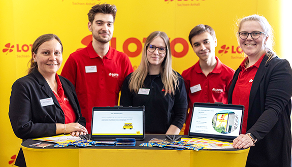 Das Messe-Team von LOTTO Sachsen-Anhalt auf der Berufsfindungsmesse der IHK in Magdeburg- Sie stehem am gelben Messestand mit dem LOTTO-Logo in Rot nebeneinander, Links Susanne Tautvetter, rechts Lisa Ritz, dazwischen drei Azubis.  