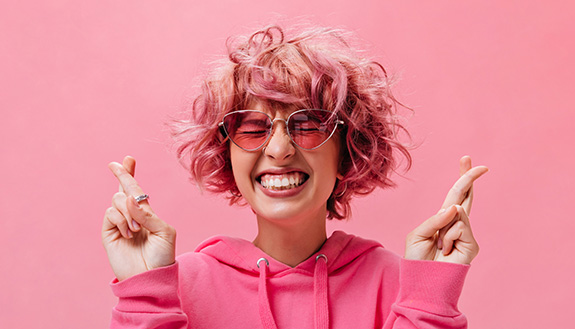 L&auml;ssige junge Frau mit pinker Sonnenbrille und Hoodie l&auml;chelt mit geschlossenen Augen und dr&uuml;ckt die Daumen