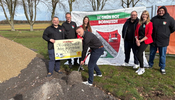 Vereinsvorstand des SV Bornstedt beim 1. Spatenstich f&uuml;r da Vereinsheim, sieben Personen stehen vor einer Fahne mit Veeinswappen auf einer Wiese und heben den ersten symbolischen Spatenstich aus. 