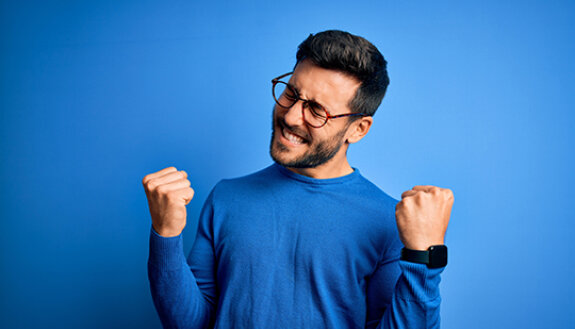 Mann jubelt mit geballten F&auml;usten im blauen Pullover vor blauem Hintergrund: Sy,mbolbild zu den Gewinnern im Spiel 77 und SUPER 6 am 7. M&auml;rz 2026. 