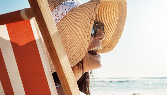 Frau mit Sonnebrille und Sonnenhut am Strand lacht freudig