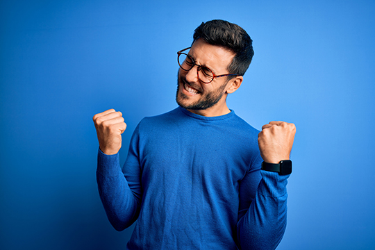 Mann jubelt mit geballten F&auml;usten im blauen Pullover vor blauem Hintergrund: Sy,mbolbild zu den Gewinnern im Spiel 77 und SUPER 6 am 7. M&auml;rz 2026. 