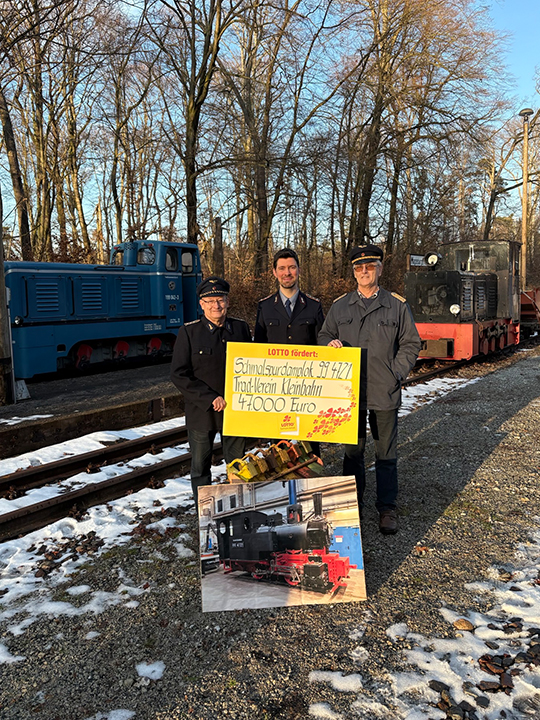Drei M&auml;nner vom Verein in historischer Uniform mit dem symbolischem Scheck von Lotto &uuml;ber 47 000 Euro und einem Foto der Lokomotive Emma.