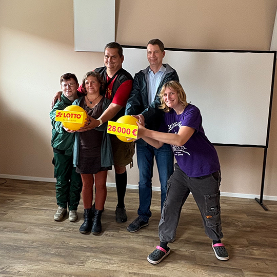 Lotto-Geschäftsführer Stefan Ebert steht mit Kristin Klatt, der Teamleiterin im Mariannenhof, und Bewohnern des Hauses im Freizeitraum mit einem symbolischen Fördercheck.