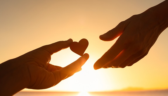 Eine Hand gibt einer anderen Hand ein Herz. Im Hintergrund ist ein Sonnenuntergang zu sehen.