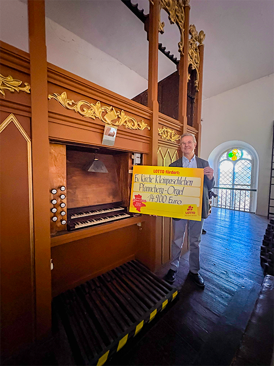 Pfarrer steht vor Orgel mit Lottoscheck in den H&auml;nden