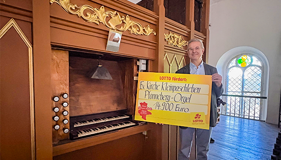 Pfarrer steht vor Orgel mit Lottoscheck in den H&auml;nden