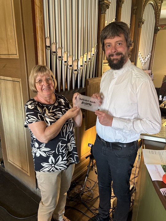 Pfarrer Karsten Höpting und Gemeindekirchenrat Birgitt Ständer Plakette „gefördert von LOTTO Sachsen-Anhalt“ vor der sanierten Knauf-Orgel in der Sankt Petruskirche in Allrode