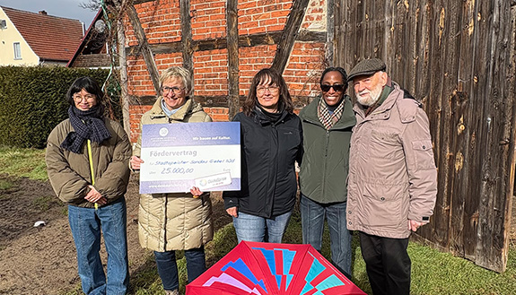 5 Personen stehen mit dem symbolischen Scheck der Gl&uuml;cksspirale und einem Regenschirm mit Gl&uuml;cksSpiralen-Logo vor dem Speicher in Sandau: V.l.n.r.: Louise Nguyen &ndash; Architektin B&auml;rbel Hornemann &ndash; Ortskuratorin Stendal der Deutschen Stiftung Denkmalschutz Birgit Pietsch &ndash; Mitarbeiterin der Stadt Sandau Vicky Germain &ndash; Eigent&uuml;merin des ehemaligen Stadtspeichers Dr. Volker Stephan &ndash; ehemaliger Ortskurator Stendal der Deutschen Stiftung Denkmalschutz