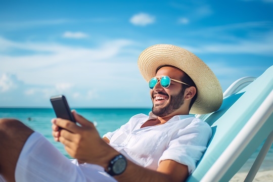 Lächelnder Mann mit Strohhut und Sonnenbrille nutzt sein Smartphone am Strand, genießt einen sonnigen Tag und entspannt am Meer