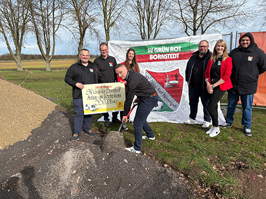 Vereinsvorstand des SV Bornstedt beim 1. Spatenstich f&uuml;r da Vereinsheim, sieben Personen stehen vor einer Fahne mit Veeinswappen auf einer Wiese und heben den ersten symbolischen Spatenstich aus. 