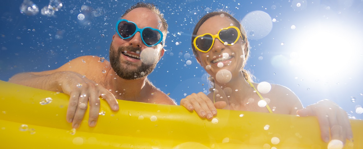 Das Bild zeigt einen fröhlichen Mann und eine Frau im Wasser, beide mit herzförmigen Sonnenbrillen und breitem Lächeln. Sie lehnen an einer gelben Luftmatratze, während Sonnenlicht und Wassertropfen eine sommerliche, unbeschwerte Stimmung erzeugen.