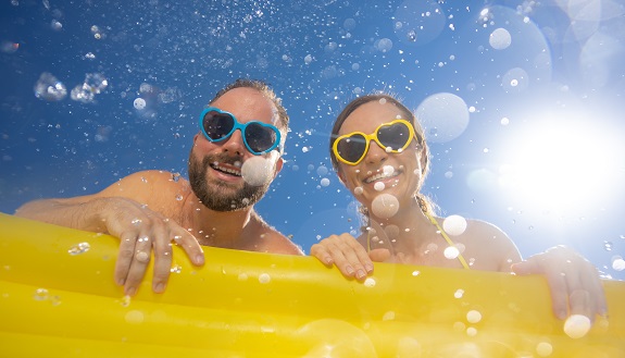 Das Bild zeigt einen fröhlichen Mann und eine Frau im Wasser, beide mit herzförmigen Sonnenbrillen und breitem Lächeln. Sie lehnen an einer gelben Luftmatratze, während Sonnenlicht und Wassertropfen eine sommerliche, unbeschwerte Stimmung erzeugen.